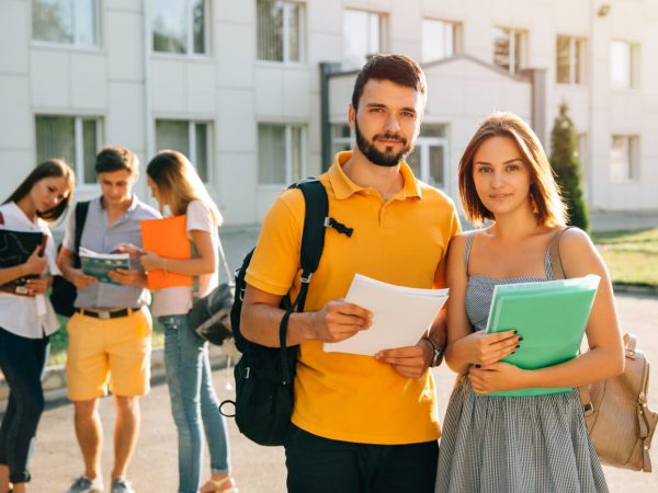 Two happy students with backpacks and books in their hands smiling at camera while standing on background of university and friends
